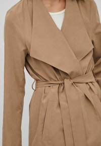 Beige omslagskappa i mjukt, strukturerat tyg med bred krage, midjebälte och två sidofickor för extra funktionalitet.