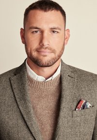 Grauer Fischgrät-Blazer über einem braunen Pullover mit geripptem Kragen, weißem Hemd darunter und einem gemusterten Taschentuch. Kurz geschnittenes Haar.