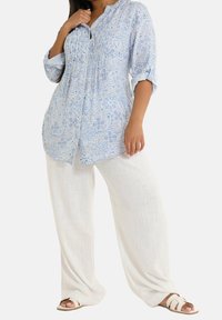 Jasnoniebieska tunika w wzór paisley z guzikami, w połączeniu z białymi, szerokimi spodniami lnianymi i beżowymi sandałami wsuwanymi. Całość z teksturowanej tkaniny.