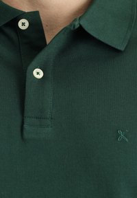Zielona koszulka polo z fakturowanej tkaniny, z trzema guzikami przy kołnierzyku, kremowymi guzikami i małym haftowanym logo na piersi.