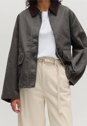 Personne portant une veste gris foncé avec un col marron par-dessus un haut blanc et un pantalon beige taille haute avec une ceinture nouée.