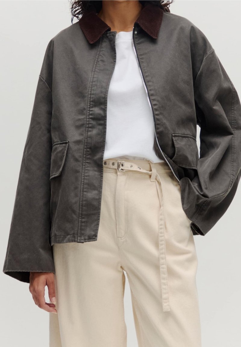 Personne portant une veste gris foncé avec un col marron par-dessus un haut blanc et un pantalon beige taille haute avec une ceinture nouée.