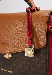Sac à main Michael Kors marron et foncé à motifs avec une bandoulière en cuir rouge et un charm cadenas doré attaché près du rabat.