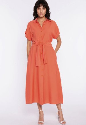 Kobieta w koralowej sukience midi z guzikami, krótkim rękawem, paskiem wiązanym w talii i białych sandałkach na wysokim obcasie z paskiem wokół kostki.