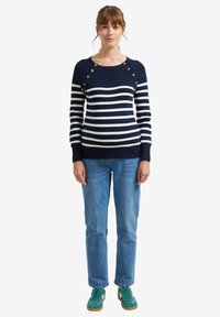 Marinegestreifter Pullover mit weißen Streifen, der über Knopfakzente an den Schultern verfügt. Kombiniert mit blauen Jeans und türkisfarbenen Sneakers.