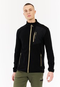 Uomo con capelli corti che indossa una giacca nera con zip beige e pantaloni verde oliva, in piedi su uno sfondo bianco.