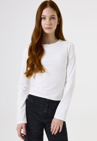 Weißes Langarmshirt mit dezenten Verzierungen, Rundhalsausschnitt, glatter Textur, kombiniert mit dunklen gemusterten Jeans.