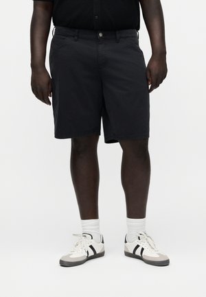 Mand iført sorte shorts, hvide crew-strømper og hvide sneakers med sorte striber, stående mod en ensfarvet baggrund.