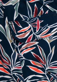 Marineblauwe stofshirt met een zak, voorzien van een meerkleurig loofpatroon in rood, groen en beige. De textuur is glad en licht van gewicht.