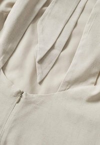 Beige kledingstuk met een diepe V-hals, bestaande uit twee overlappende stofstukken en een ritsluiting aan de zijkant, gemaakt van textuurmateriaal.