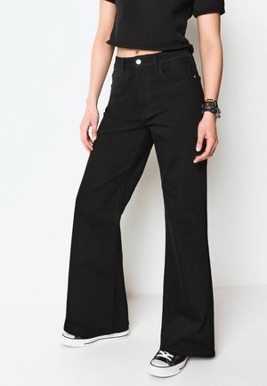 Pantaloni flared din denim negru cu un design cu cinci buzunare, mulati pe talie și șolduri, având fermoar și închidere cu button, purtați cu adidași negri.