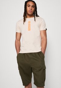 Beige bomulds t-shirt med et orange vertikalt "ALPHA" logo på brystet, parret med grønne cargo shorts med sidelommer.