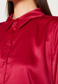 Czerwona satynowa bluzka z klasycznym kołnierzykiem, pięcioma guzikami i gładką teksturą. Tkanina odbija światło, podkreślając jej błyszczące wykończenie.