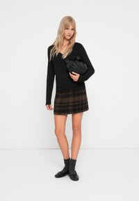 Schwarzes Langarmoberteil, karierter Minirock in Brauntönen, schwarze Handtasche, graue Socken und schwarze Ballerinas. Schlichter weißer Hintergrund.