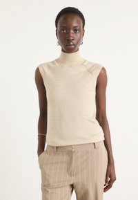 Bež brezrokavni turtleneck top z oprijetim silhueto, narejen iz mehkega materiala, z diskretno teksturo in rebrastim ovratnikom.