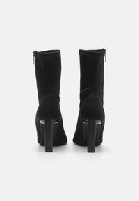 Botas de tobillo negras hechas de un tejido elástico con una textura suave, que cuentan con un alto tacón grueso y un detalle de cremallera lateral.