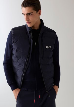 Jeune homme portant un gilet matelassé bleu marine sur un pull zippé, debout les mains dans les poches, regardant sur le côté sur un fond neutre.