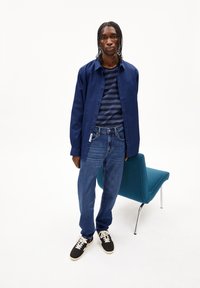 Navy knoopshirt over een marineblauwe en grijs gestreepte T-shirt, gecombineerd met blauwe spijkerbroek en zwarte sneakers met witte veters.