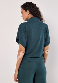 Teal stickad topp med hög krage, korta ärmar och avslappnad passform; matchas med byxor i samma färg. Mjuk textur, minimal detaljering och inga synliga mönster.