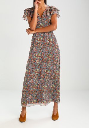 Femme portant une longue robe fleurie avec des manches courtes à volants et des chaussures à talons marron, debout avec une main près du visage et l'autre croisée.