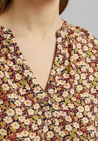 Blusa floreale con scollatura a V profonda, caratterizzata da piccoli fiori bianchi e foglie verdi su uno sfondo nero, con dettagli di arricciatura al colletto.