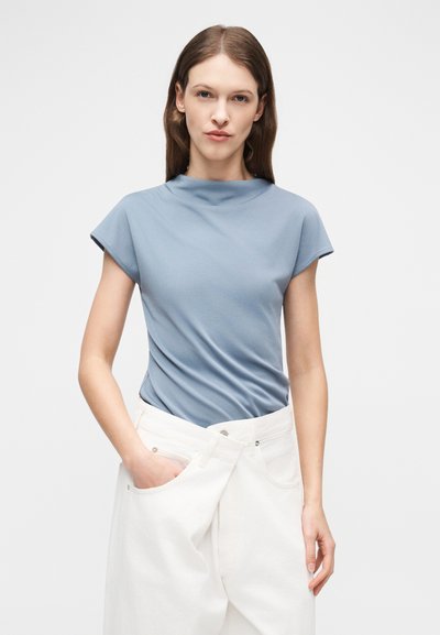 Femme aux cheveux bruns lisses portant un haut bleu clair à manches courtes et un pantalon blanc ample, une main dans la poche, sur fond uni.