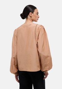 Blouse oversize beige avec des manches bouffantes, présentant des plis détaillés aux poignets et un col rond. Tissu lisse et silhouette décontractée.