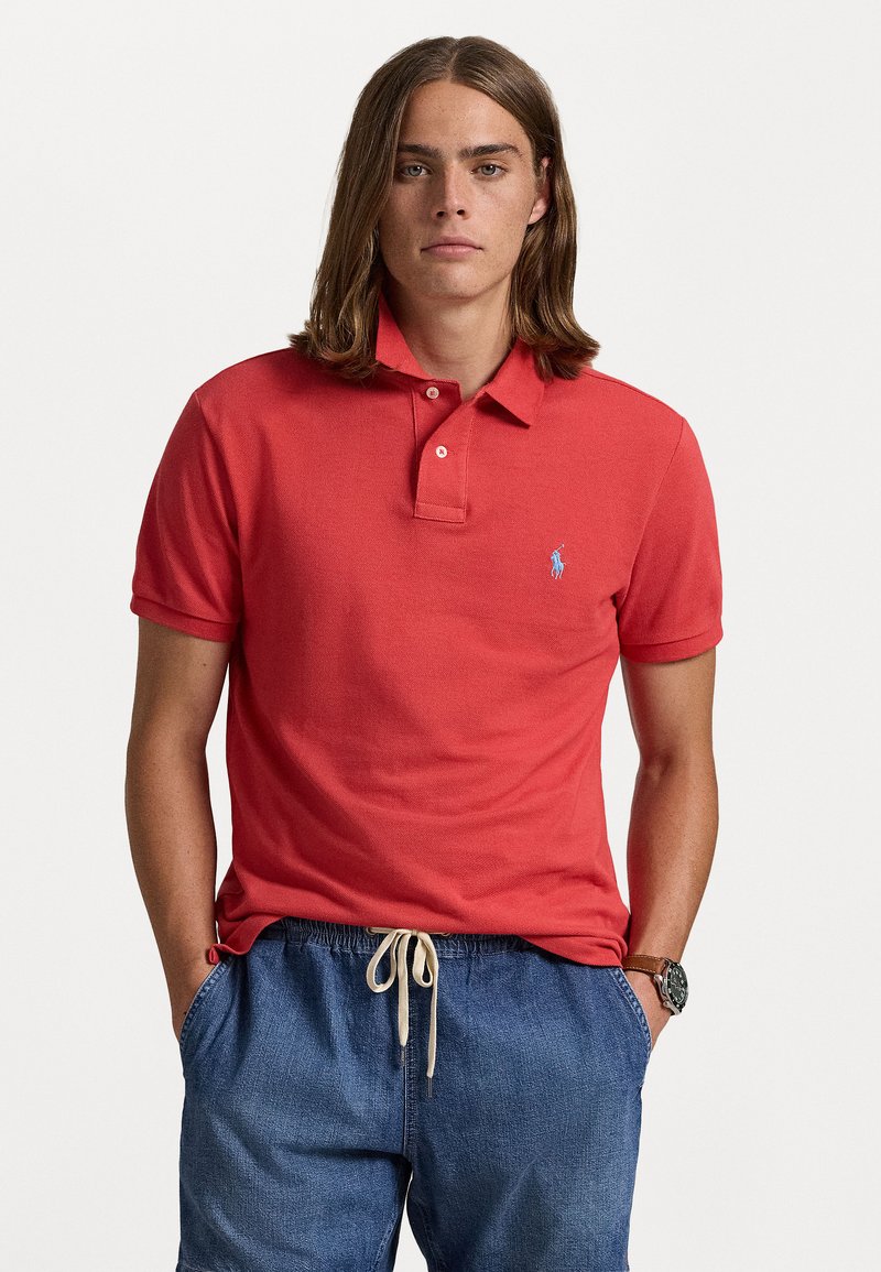 Röd polo t-shirt i texturerad tyg med tre-knapps knäppning och liten blå logotyp på vänster bröst, i kombination med denimshorts.