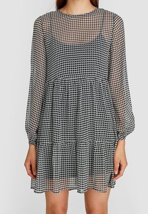 Vrouw draagt een zwart-witte doorzichtige jurk met pied-de-poulle patroon, met lange mouwen en een getailleerde taille.