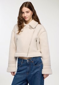 Giacca in shearling beige tagliata con ampio colletto, cerniere in argento e texture morbida. Abbinata a jeans in denim blu.