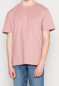 T-shirt in cotone rosa con scollatura tonda e maniche corte. Presenta una vestibilità comoda e una texture liscia, abbinata a jeans in denim blu chiaro.