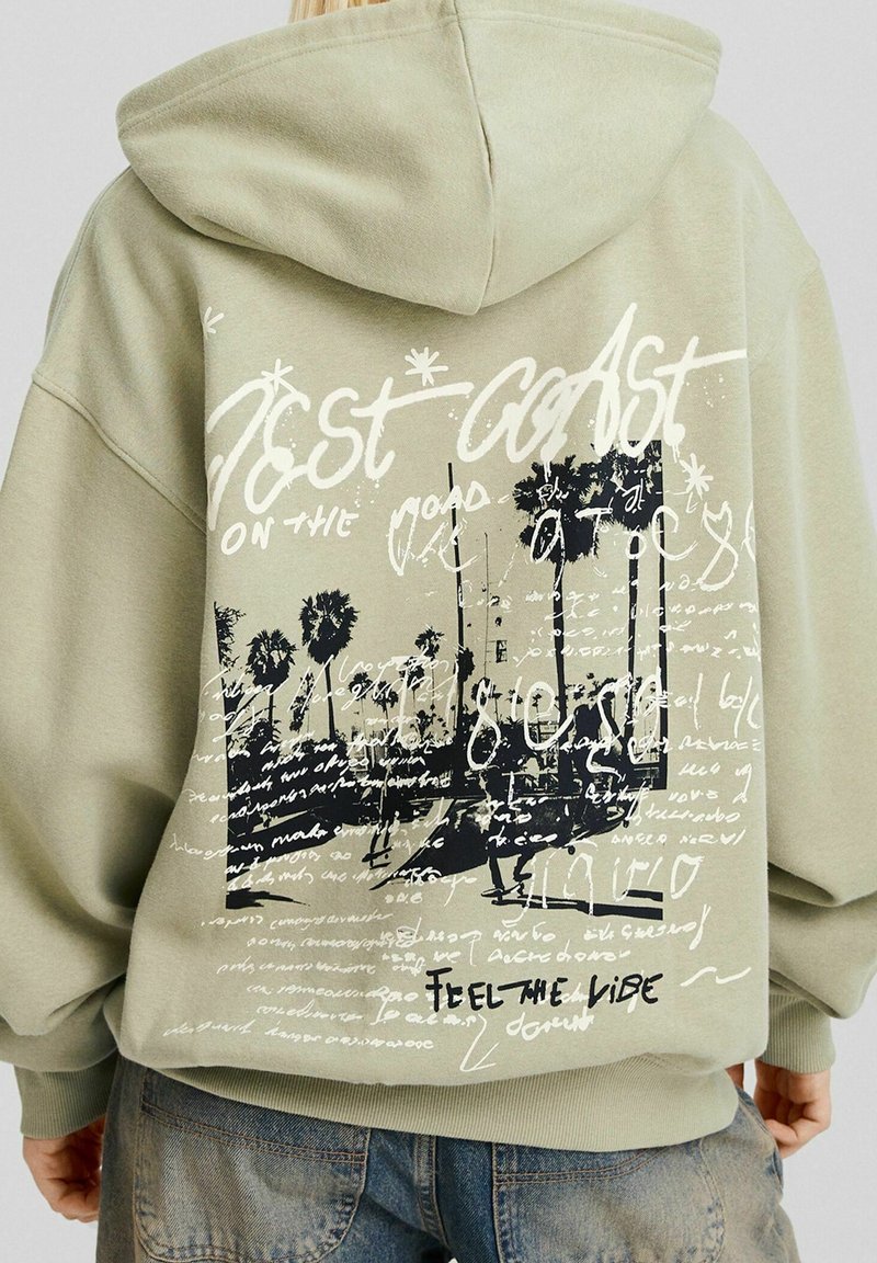 Olivengroene hoodie met een grafische print van palmbomen, handgeschreven tekst en een capuchon. Tekst bevat "West Coast" en "Feel the Vibe."