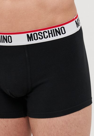 Mustat boxeri alushousut, jotka on valmistettu pehmeästä kankaasta. Niissä on valkoinen vyötärönauha punaisilla yksityiskohdilla ja musta "MOSCHINO" -logo-teksti.
