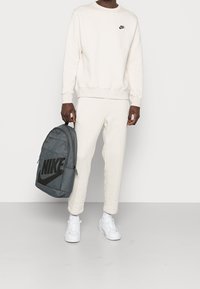 Svetle béžová Nike mikina a tepláky, doplněné bílými teniskami. Šedý Nike batoh má velké černé logo.