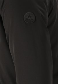 Schwarze Jacke aus strukturiertem Stoff mit einem runden, erhabenen Logo-Patch auf dem Oberarm, das Berg- und Wellenmotive zeigt.