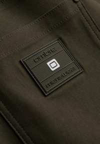 Olivgrön tyg med en texturerad, svart gummilapp med upphöjda bokstäver och en vit fyrkantig logotyp, sydd på plagget.