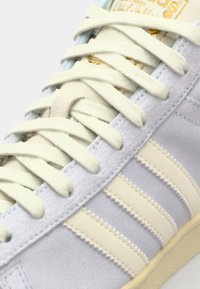 Ljusgrå mockasneakers med krämfärgade ränder, platta snören och en strukturerad sula. Har en guldfärgad logotyp på tungan och sömnadsdetaljer.