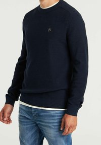 Sweatshirt en maille bleu marine avec un col rond et des poignets côtelés. Il présente un petit logo noir sur la poitrine et une chemise blanche superposée en dessous.