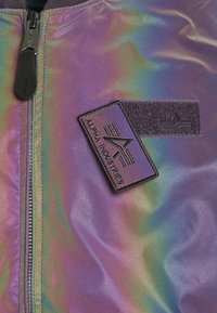 Irydescentna kurtka bombowiec o gładkiej teksturze, z zamkiem błyskawicznym i czarną naszywką z logo "ALPHA INDUSTRIES".