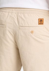 Pantaloni corti in cotone beige con fascia elastica in vita, due tasche posteriori, chiusura con bottone e una patta con il logo Timberland sul retro destro.