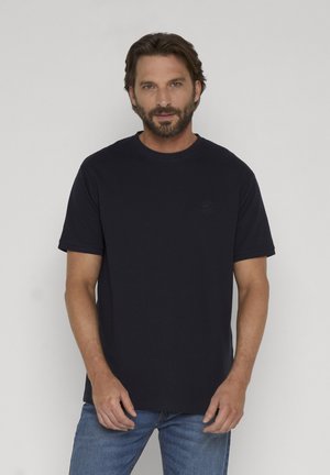 Homme aux cheveux bruns courts et barbe, portant un T-shirt noir uni à manches courtes et un jean bleu, debout devant un fond clair.