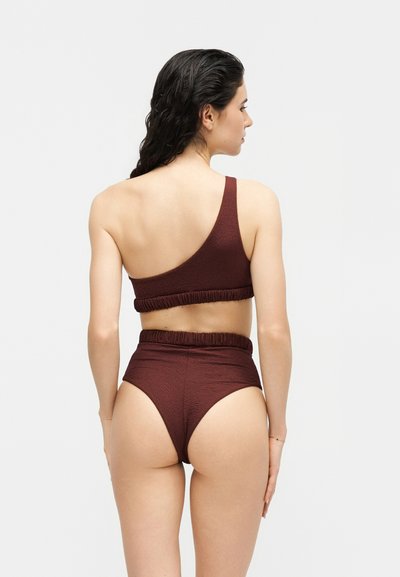 Femme aux longs cheveux noirs portant un haut de bikini marron texturé à une épaule et un bas taille haute, debout de dos face à la caméra.