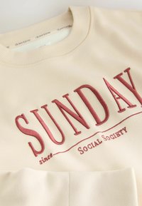 Beige sweatshirt med stort broderet "SUNDAY" i mørkerød, med "since" og teksten "Social Society" nedenfor. Blød tekstur og ribbet halsudskæring.