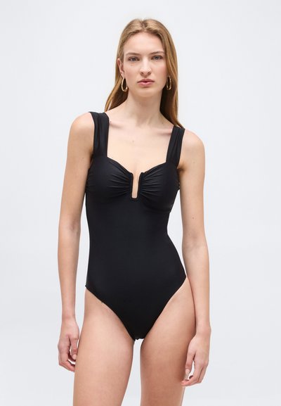 Jeune femme aux longs cheveux blonds portant des boucles d'oreilles en or en forme d'anneau et un maillot de bain noir sans manches avec un décolleté carré et un buste froncé.