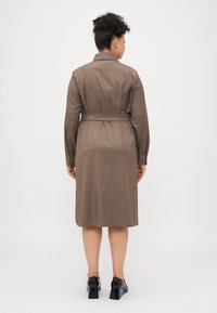 Barna övvel ellátott ruha gallérral és hosszú ujjakkal, texturált anyagból készült, középhosszú szabású. Fekete magassarkú cipővel viselve.