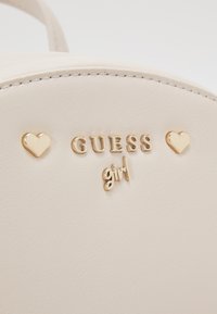 Sac en simili cuir crème avec marquage doré "GUESS girl" et deux accents en forme de cœur sur le devant, présentant une texture lisse et des lignes épurées.