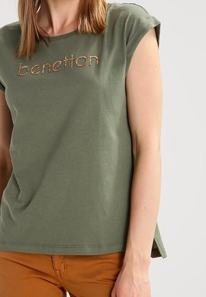 Kobieta w oliwkowej koszulce Benetton z podwiniętymi rękawami i spalonych pomarańczowych spodniach, przycięta na wysokości ramion i talii.