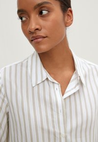 Camicia bianca con bottoni, caratterizzata da sottili righe beige, colletto e vestibilità informale. Il tessuto sembra leggero e leggermente strutturato.