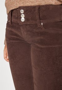 Hnědé kordové kalhoty s vertikálním žebrováním, střih s přiléhavým designem a třemi stříbrnými knoflíky na předním pásu.