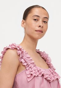 Rosa ermeløs bluse med volangkant langs halslinjen og skulderstropper, med en glatt stofftekstur.